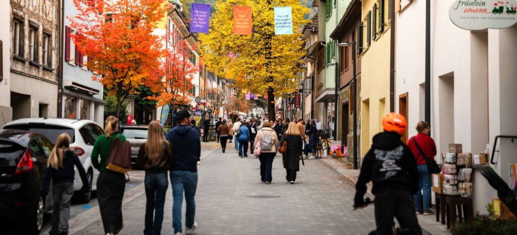 Herbstliche Gasse mit bunten Bäumen und vielen Menschen, ein Radfahrer fährt vorbei.